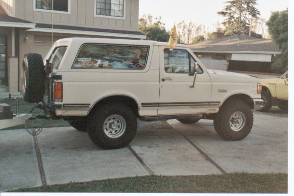White Ford Bronco, side view, with spare tire on rear door, lifted suspension, and a yellow flag on the roof