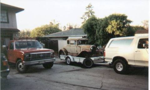 1990 Ford Bronco - A collection of vehicles, including a pickup truck, a Bronco, and an antique car on a trailer, are parked in a residential driveway.