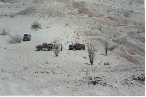 1990 Ford Bronco - Three SUVs are parked in a sandy desert area with sparse vegetation and rolling hills in the background.