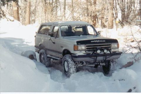 1990 Ford Bronco - A 1990 Ford Bronco navigates a snowy path, its tires deep in the snow, surrounded by trees.