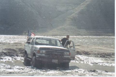 1990 Ford Bronco - Ford Bronco stuck in mud, driver standing by open door.