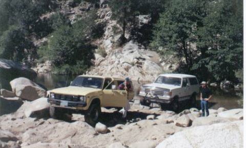 1990 Ford Bronco - Two SUVs are parked on a rocky shore next to a body of water with trees in the background.