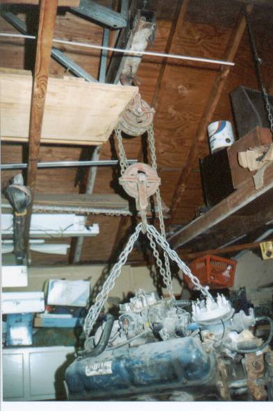 1966 Ford Bronco - An engine is hoisted by a chain system in a cluttered garage space.