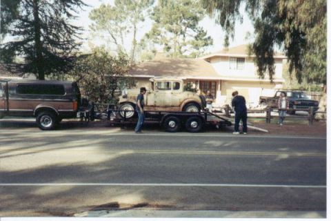 1966 Ford Bronco - Street-side scene of a rusty 1930s Ford Model A coupe project loaded on a dual-axle flatbed trailer behind a two-tone brown and black van,…