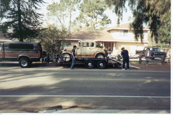 Rusty 1930s Ford Model A coupe on flatbed trailer behind a brown van at curbside