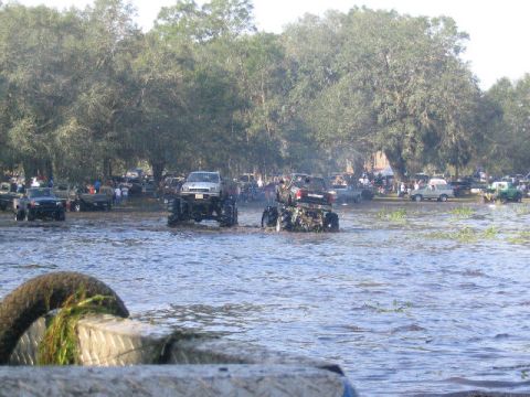 1993 Ford Bronco - Multiple monster trucks navigate a watery terrain with a crowd gathered nearby.