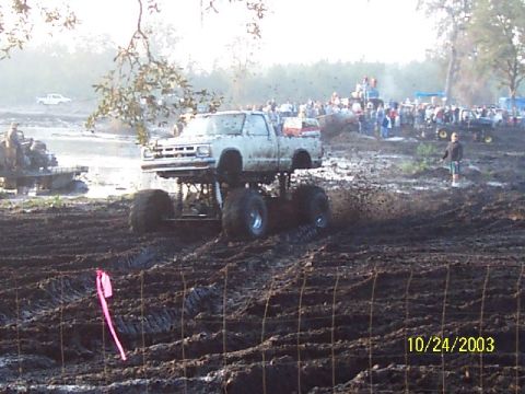 1993 Ford Bronco - A lifted pickup truck navigates a muddy track during an event, with spectators gathered to watch.