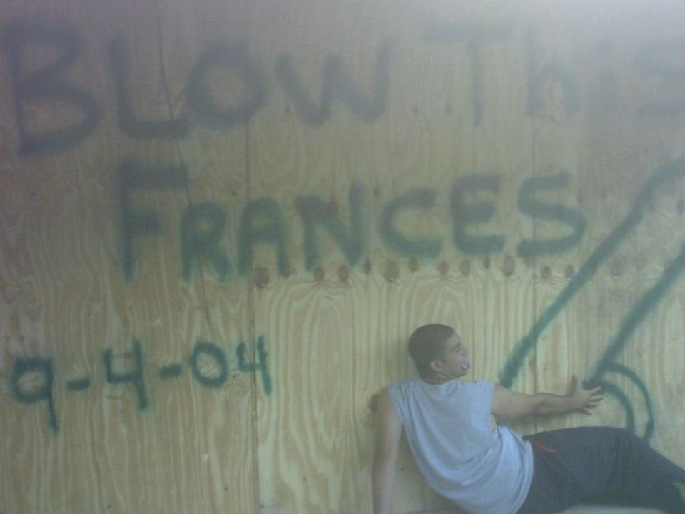 Man in grey t-shirt leaning against a plywood wall with green graffiti that reads "BLOW TH FRANCES 9-4-04"