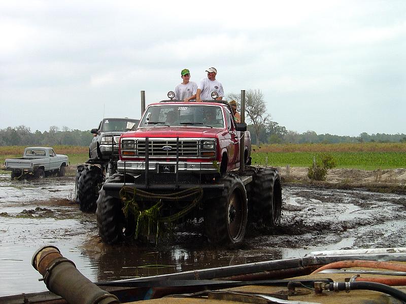 Modified red Ford pickup truck with large tires in a muddy field, two people on top. Another truck is behind it