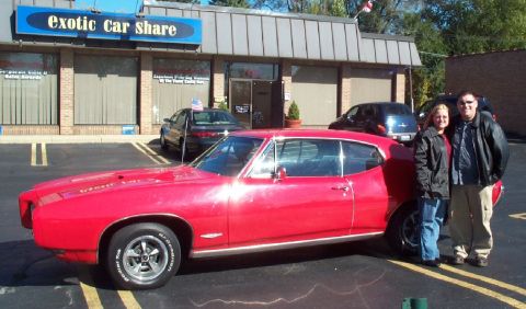 1985 Ford Crown Victoria LTD - A red 1967 Pontiac GTO coupe is parked outside Exotic Car Share, with two people posing next to it.
