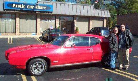 1985 Ford Crown Victoria LTD - A bright red 1969 Pontiac GTO is parked outside an Exotic Car Share business with two people standing next to it.