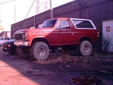 1986 Ford Bronco - A red Ford Bronco with oversized tires sits parked outside a commercial building.
