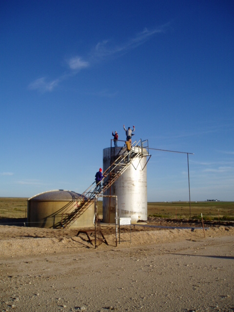 1990 Ford Bronco - People stand on the stairs and platform of tall industrial tanks under a blue sky.
