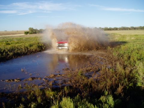 1990 Ford Bronco - A red truck creates a massive mud splash as it drives through a water-filled ditch in a grassy field.