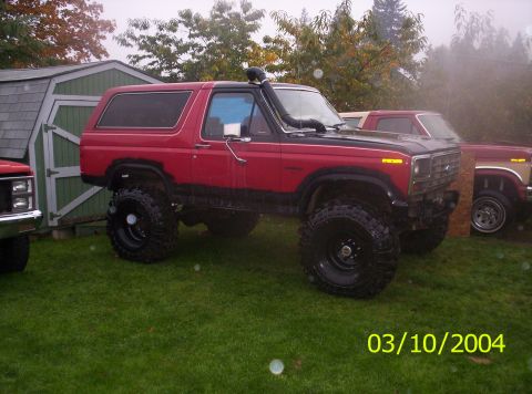 1983 Ford Bronco - Modified red Ford Bronco with snorkel and large tires sits on a grassy lawn near a shed and another truck.