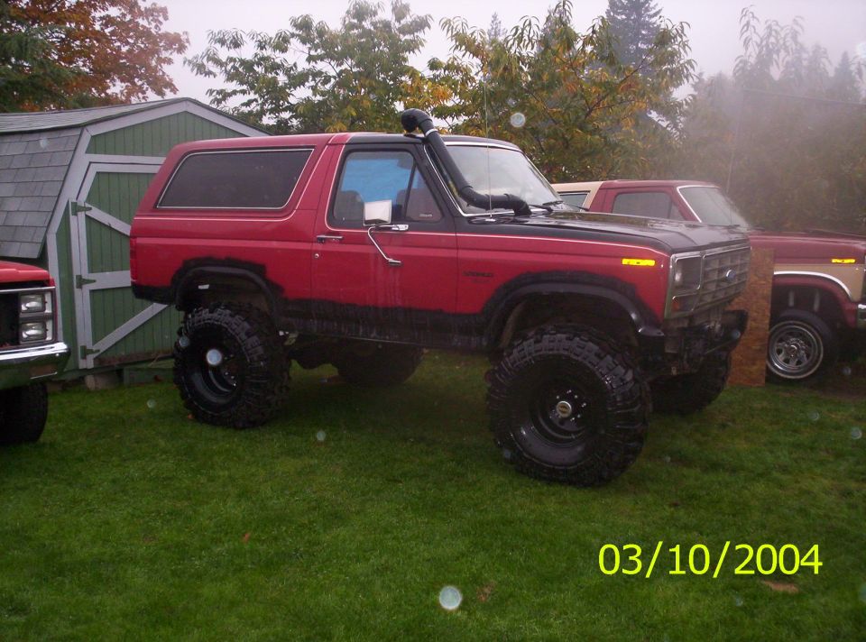 Lifted red Ford Bronco with snorkel and off-road tires parked on grass next to a shed. Another Ford truck is visible behind it