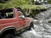 1983 Ford Bronco - Very small, low-resolution thumbnail of a red full-size Ford Bronco-style SUV with a white roof fording a rocky creek, seen from the front…