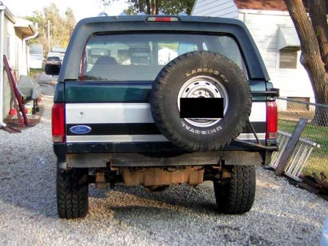 1995 Ford Bronco - The rear of a dark green Ford Bronco is shown with a spare tire and a ladder leaning against a fence nearby.