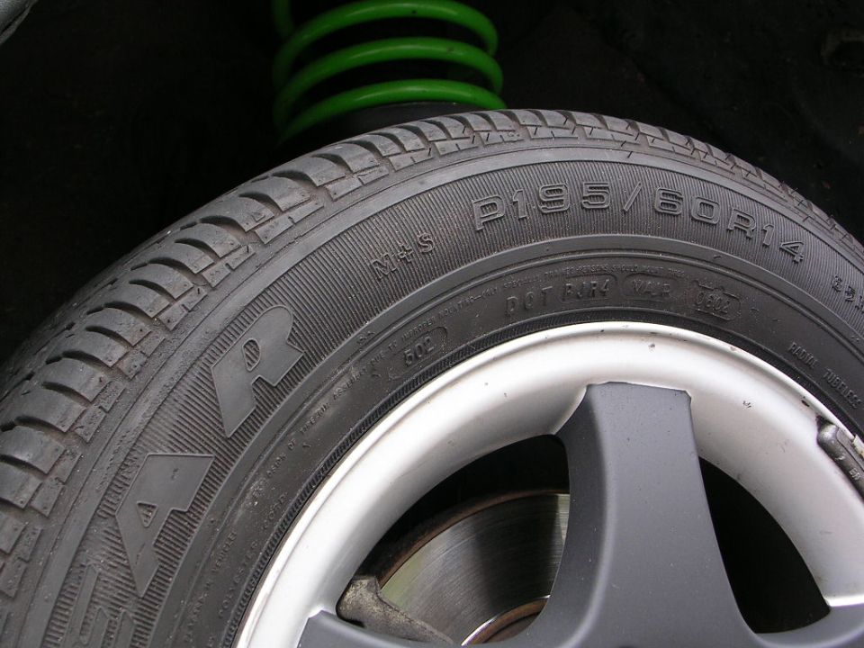 Close-up of a car tire with P195/60R14 markings and a green coil spring in the background