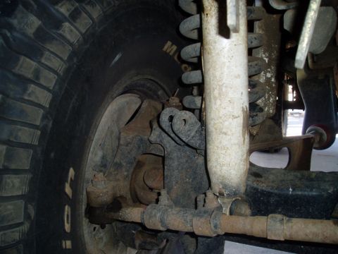 1990 Ford Bronco - Close-up under a lifted 4x4 showing the front solid axle knuckle, coil spring, white-bodied shock absorber and a tie rod with adjusting sle…