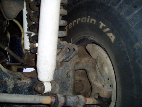 1990 Ford Bronco - Close-up under a truck's front end showing a white-bodied shock absorber and blue-painted coil spring above a dusty solid-axle knuckle, wit…