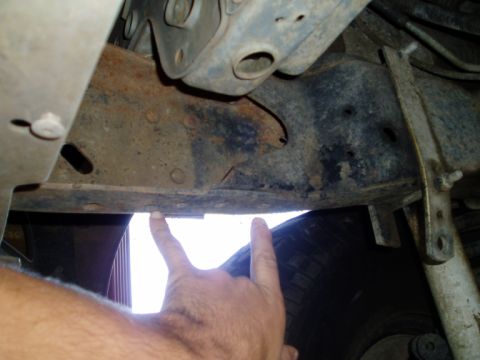 1990 Ford Bronco - An arm reaches under a truck and two fingers point at a pair of holes in the rusty boxed frame rail, indicating mounting points beneath the…