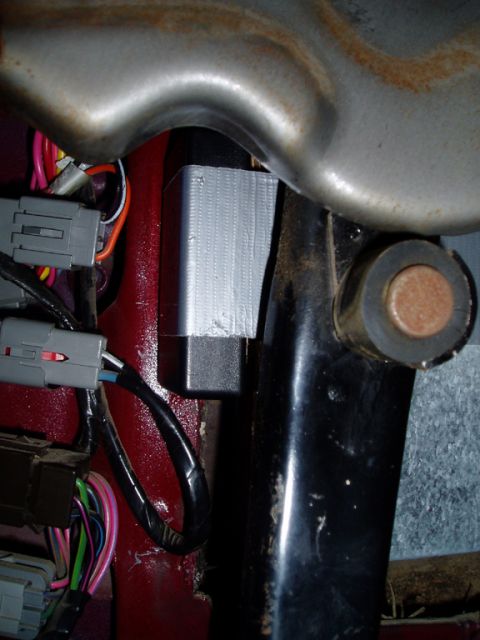 1990 Ford Bronco - Tight engine-bay shot of a black electronic module wrapped in silver duct tape, tucked between the red inner fender panel and a black metal…