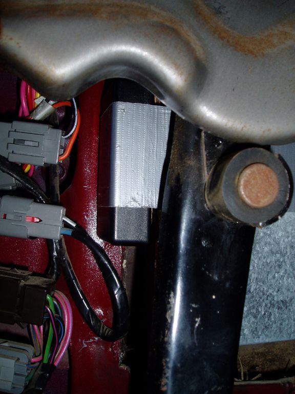Duct-taped black module wedged beside a red inner fender and exhaust pipe with wiring