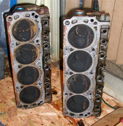 1990 Ford Bronco - Two rusty cast-iron V8 cylinder heads sitting combustion-chamber up on a plywood surface in a workshop.