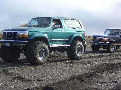 1996 Ford Bronco - A lifted green 1996 Ford Bronco is parked next to a dark blue Ford pickup truck on a dirt road.