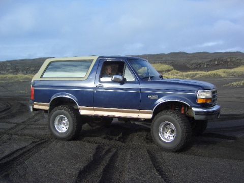 1996 Ford Bronco - A blue 1996 Ford Bronco with a tan top drives on a black sand beach under a cloudy sky.