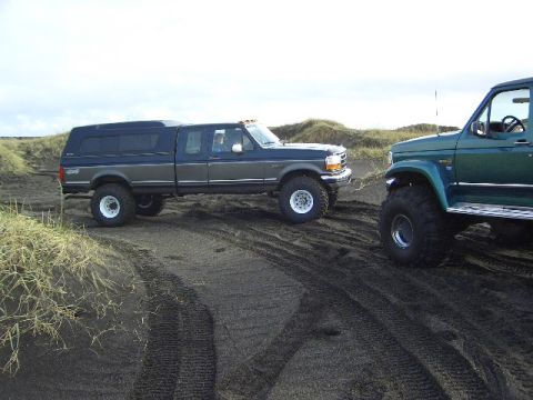1996 Ford Bronco - Two lifted pickup trucks are parked on a sandy surface, with one truck in a two-tone blue and gray color scheme and the other in green.