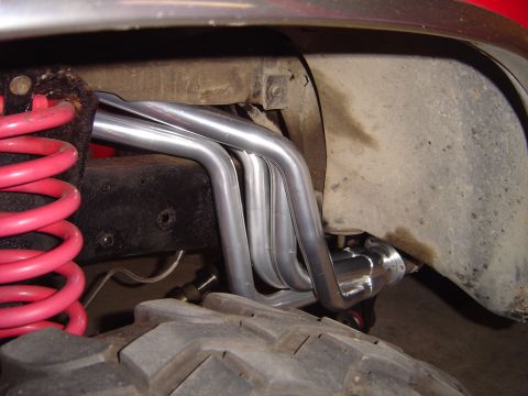 1978 Ford Bronco - Wheel-well view of unpainted stainless long-tube headers routed down past the frame rail on a lifted red truck, with a red coil spring at l…
