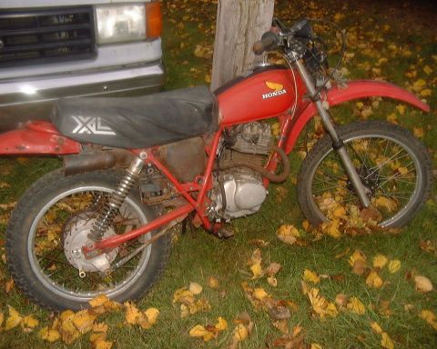 1985 Ford F-150 - A red Honda XL motorcycle is parked on grass with fallen leaves, next to the front of a silver truck.