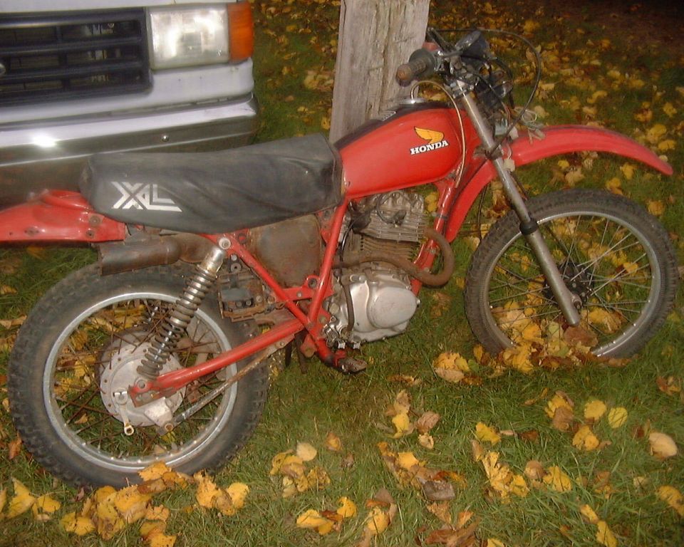 Red vintage Honda XL dirt bike on a leaf-covered lawn beside a white pickup, right-side view