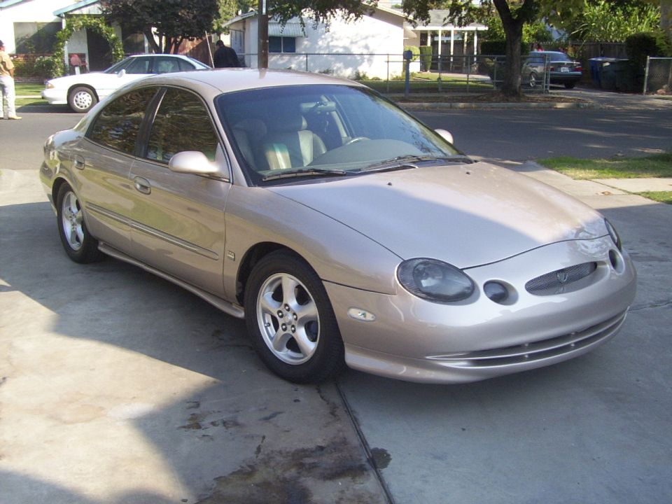 Tan sedan parked on a driveway, front quarter view. The car has tinted windows and alloy wheels