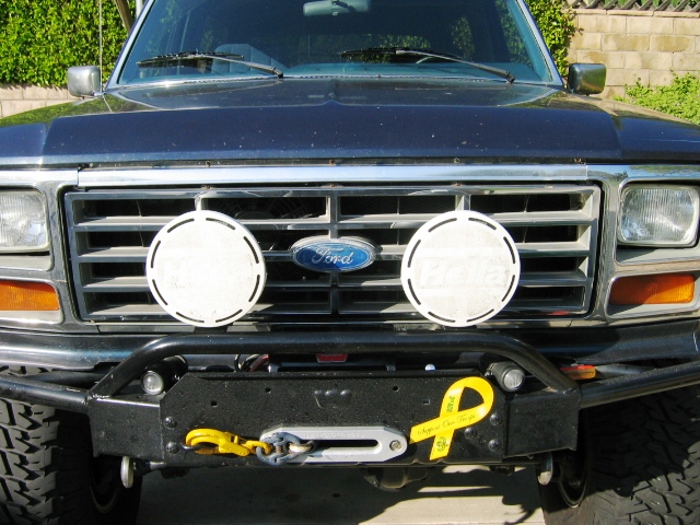 Front of dark Ford Bronco with grille, two round driving lights, winch bumper and yellow ribbon.
