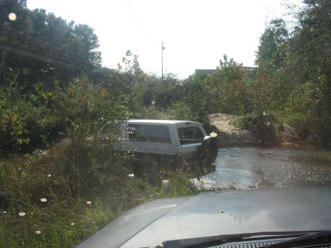 1988 Ford Bronco - A silver pickup truck with a camper shell is stuck in shallow water surrounded by vegetation, with another vehicle visible in the foregroun…