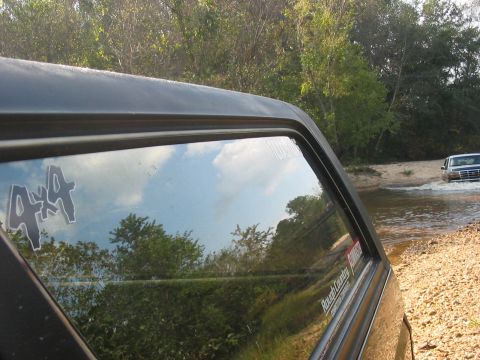 1988 Ford Bronco - A black SUV with a 4x4 sticker navigates a shallow water crossing, with a white SUV visible in the distance.