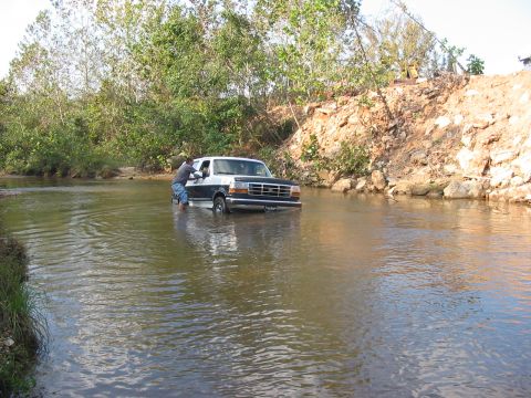 1988 Ford Bronco - A person attempts to push a white and black SUV that is stuck in a shallow river.