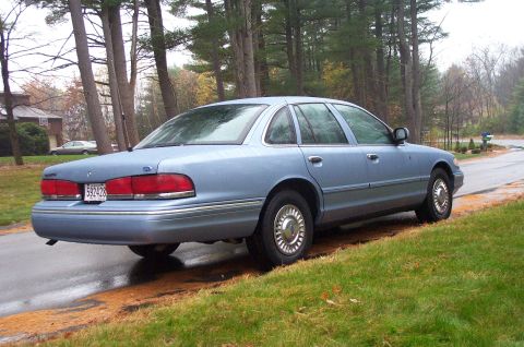 1991 Ford Crown Victoria LTD - A light blue sedan is parked on the side of a road on a cloudy day, with trees and a house visible in the background.