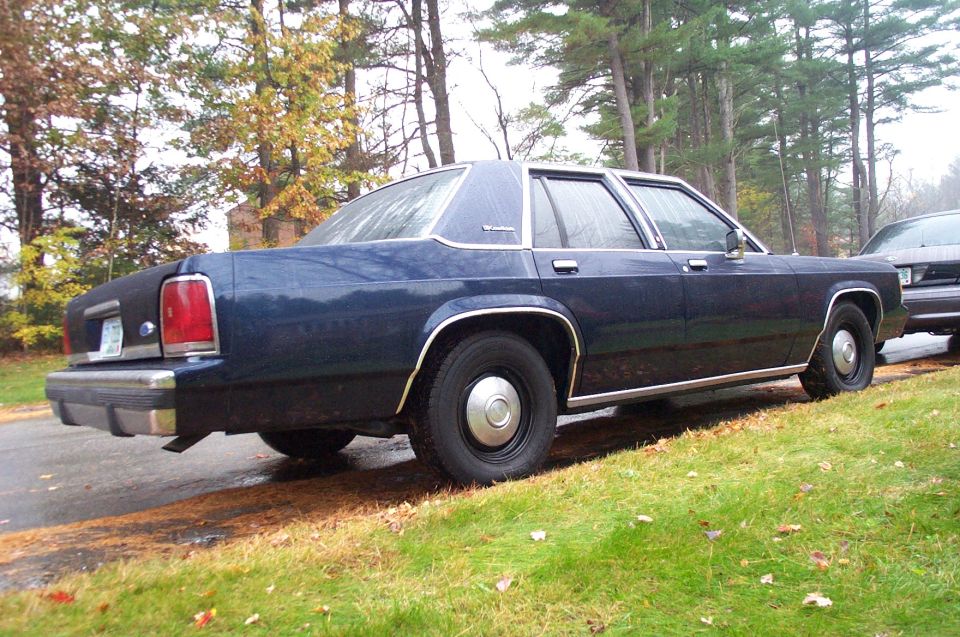 Dark blue sedan parked on a grassy shoulder, side view. Another vehicle is partially visible to the right