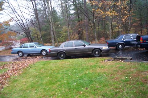 1991 Ford Crown Victoria LTD - Three sedans and a pickup truck are parked on a wet driveway during autumn.