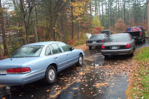 1991 Ford Crown Victoria LTD - Several vehicles are parked on a wet, leaf-covered driveway in a wooded area during autumn.