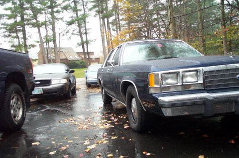 1991 Ford Crown Victoria LTD - Multiple vehicles are parked on a wet, leaf-covered driveway in front of a house.