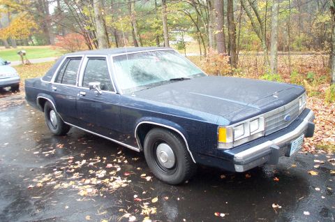 1991 Ford Crown Victoria LTD - A dark blue Ford Crown Victoria sits parked on a wet driveway surrounded by autumn leaves.
