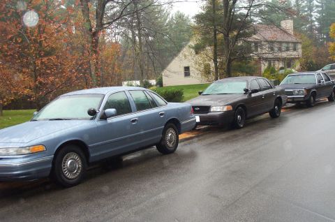 1991 Ford Crown Victoria LTD - A line of police cars are parked on a street on a rainy day, with autumn trees in the background.