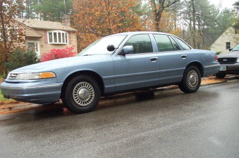 1991 Ford Crown Victoria LTD - A blue 1991 Ford Crown Victoria LTD sedan is parked on a wet street, with another vehicle visible behind it.