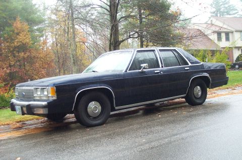 1991 Ford Crown Victoria LTD - A dark blue sedan is parked on the side of a wet road during a rainy day, with autumn foliage visible.