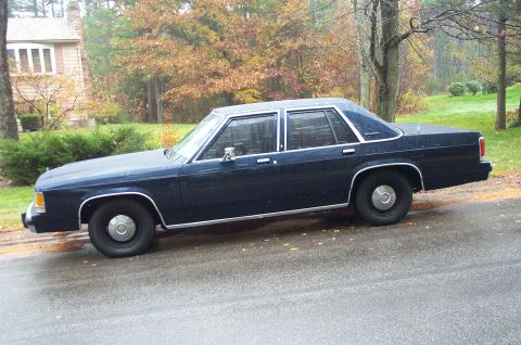 1991 Ford Crown Victoria LTD - A dark blue sedan with chrome trim is parked on a wet road, with a house and trees in the background.